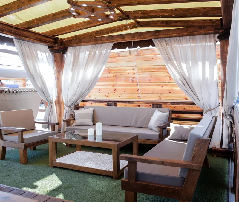 Terasă acoperită din lemn cu draperii crem strânse pe stâlpi, canapele și fotolii cu perne, perete din lemn și gazon artificial — LEMNER.MD