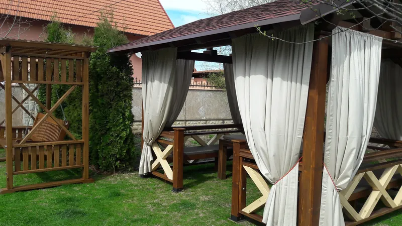 Foișor din lemn pe gazon cu draperii bej deschise, balustradă în X și bănci interioare, lângă un leagăn din lemn — LEMNER.MD