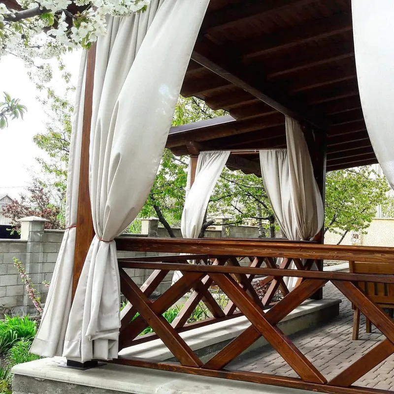 Foișor din lemn cu draperii crem pe lateral și balustradă în X, cu masă și bănci din lemn lângă un gard de piatră — LEMNER.MD