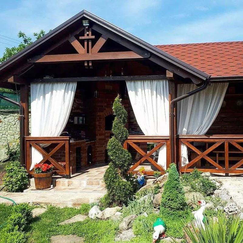 Terasă acoperită lângă casă cu acoperiș roșu în două ape, draperii albe între stâlpi și balustradă în X, cu grătar din piatră — LEMNER.MD