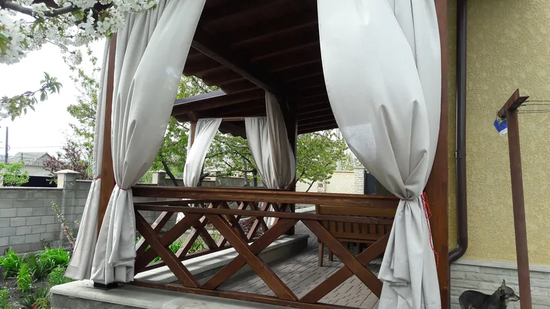 Foișor din lemn închis cu draperii albe și prelate transparente, cu balustradă în X, pe terasă pavată lângă casă — LEMNER.MD