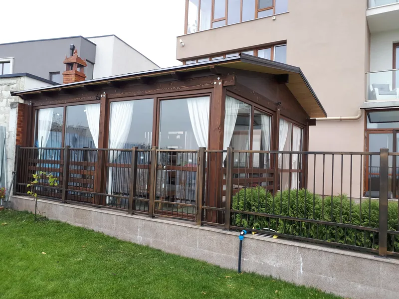 Foișor închis din lemn cu pereți de sticlă și draperii albe, parapet din scânduri, lângă bloc, pe gazon — LEMNER.MD