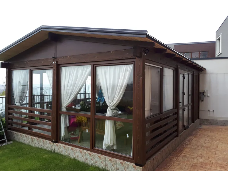 Foișor închis din lemn în nuanță de nuc, tip verandă, cu pereți de sticlă și draperii albe, acoperiș în patru ape, pe gazon — LEMNER.MD