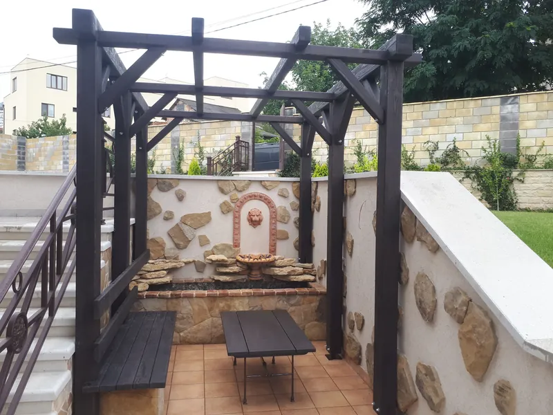 Pergolă din lemn vopsit negru cu bancă și masă, lângă un perete placat cu piatră și o fântână decorativă, în grădină — LEMNER.MD