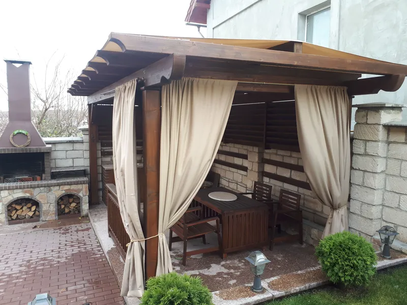 Foișor din lemn cu draperii bej, masă și scaune, lângă o zonă de grătar din cărămidă — LEMNER.MD