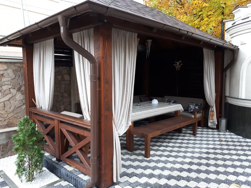 Foișor din lemn cu draperii bej, masă și bancă, pe o terasă cu pavaj decorativ, lângă casă — LEMNER.MD