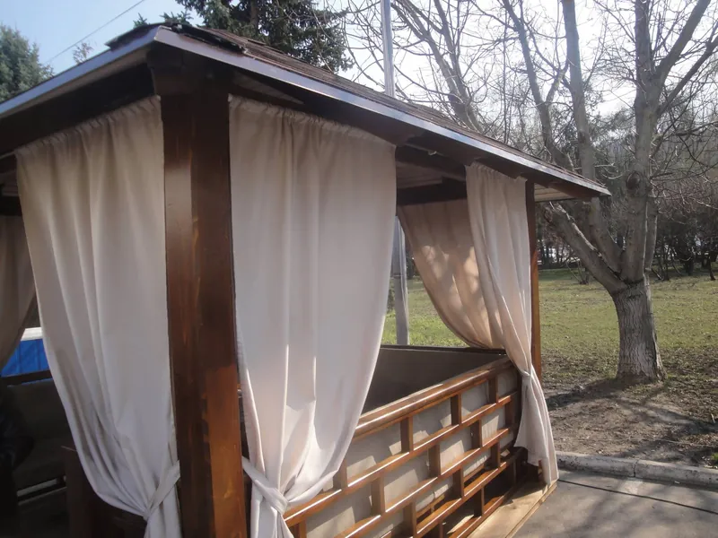 Foișor din lemn cu draperii bej și balustradă din scânduri, pe gazon primăvara — LEMNER.MD