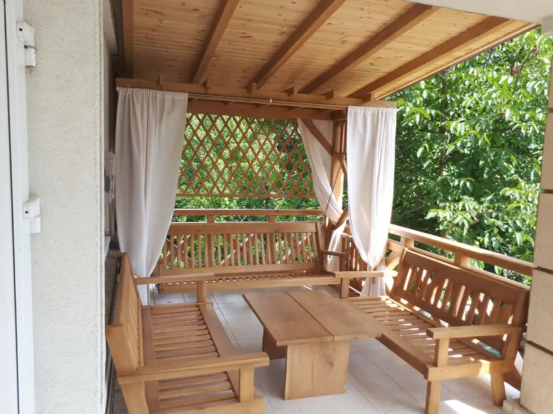 Terasă acoperită din lemn cu draperii bej, balustradă cu zăbrele și mobilier masiv din lemn, cu vedere spre grădină — LEMNER.MD