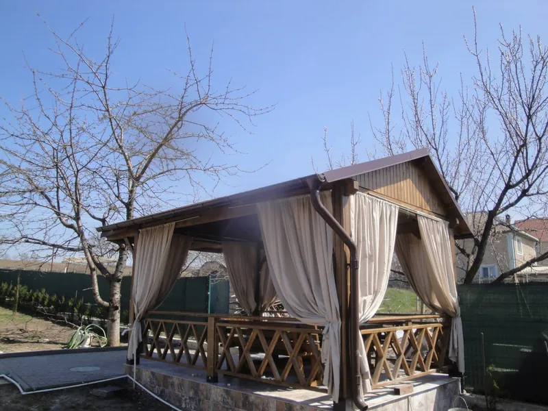 Foișor din lemn cu acoperiș în două ape, draperii bej și balustradă cu zăbrele, în grădină primăvara — LEMNER.MD