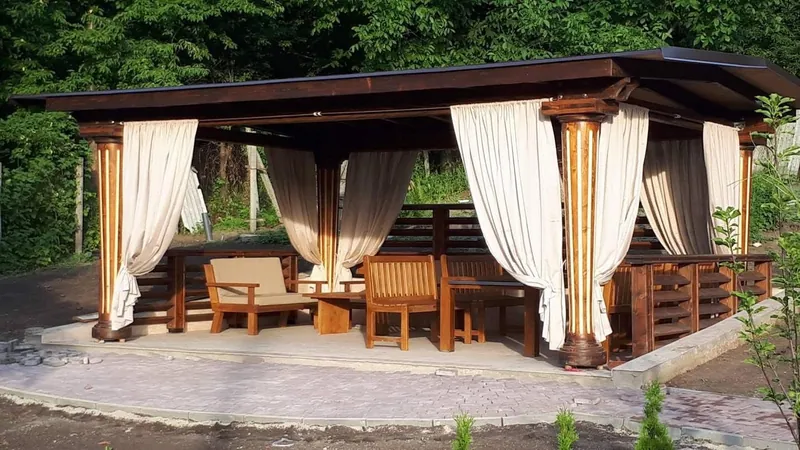 Foișor mare din lemn cu draperii bej, mobilier de relaxare și stâlpi canelați, la apus lângă pădure — LEMNER.MD