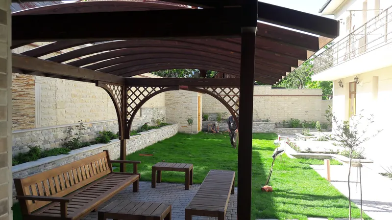 Pergolă din lemn cu arcade decorative peste o zonă de relaxare cu bănci, într-o curte amenajată — LEMNER.MD