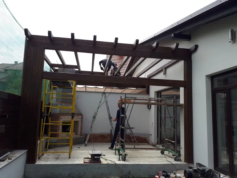 Structura din grinzi de lemn a unei copertine în construcție, cu muncitor pe schelă, lângă casă — LEMNER.MD