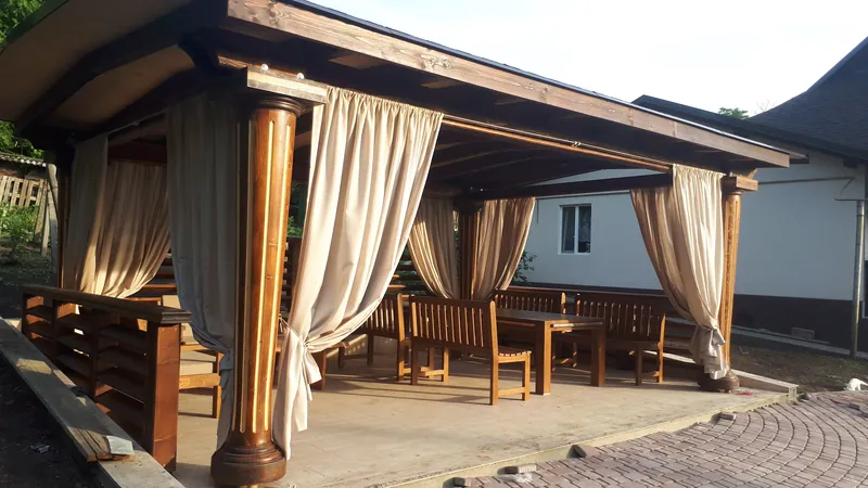 Foișor mare din lemn cu draperii bej, mese și bănci, pe o terasă pavată, la apus — LEMNER.MD