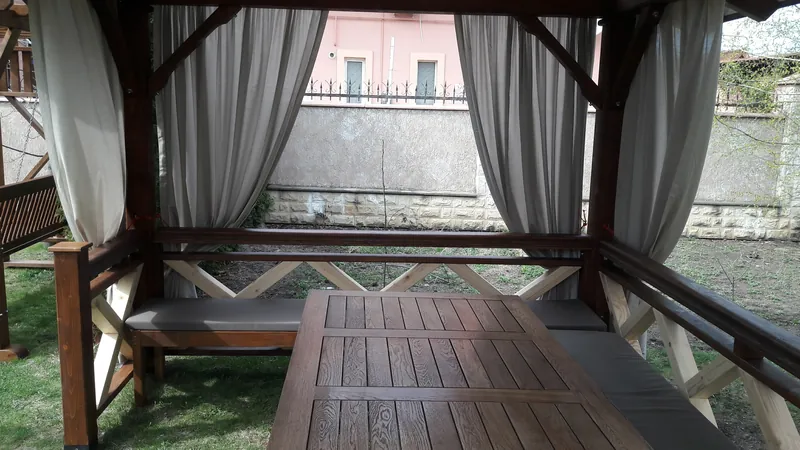 Masă din lemn cu bancă tapițată sub un foișor cu draperii albe, în grădină — LEMNER.MD