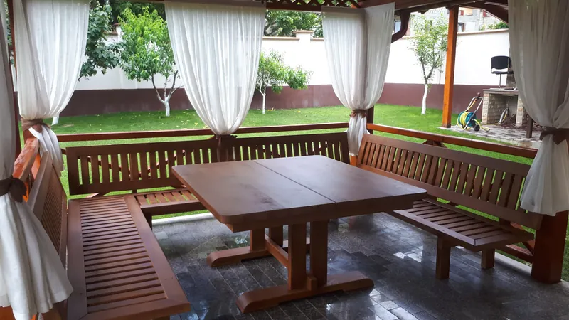 Set masă cu bănci din lemn sub un foișor cu draperii albe, cu vedere spre grădină — LEMNER.MD