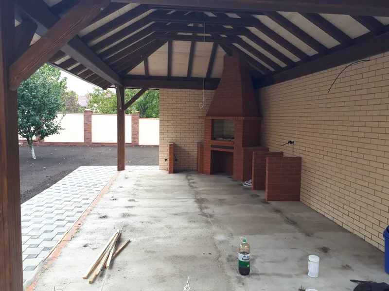 Zonă de grătar din lemn în construcție, cu acoperiș în patru ape și grătar din cărămidă lângă un perete din cărămidă — LEMNER.MD