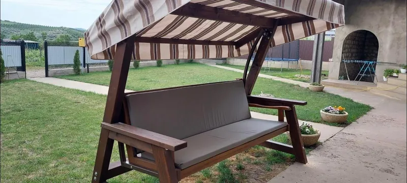 Leagăn din lemn cu copertină textilă în dungi și pernă gri, pe peluză cu priveliște deschisă — LEMNER.MD