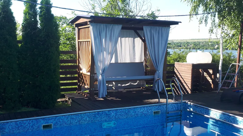 Leagăn din lemn cu acoperiș și perdele albe, amplasat pe terasă lângă piscină — LEMNER.MD