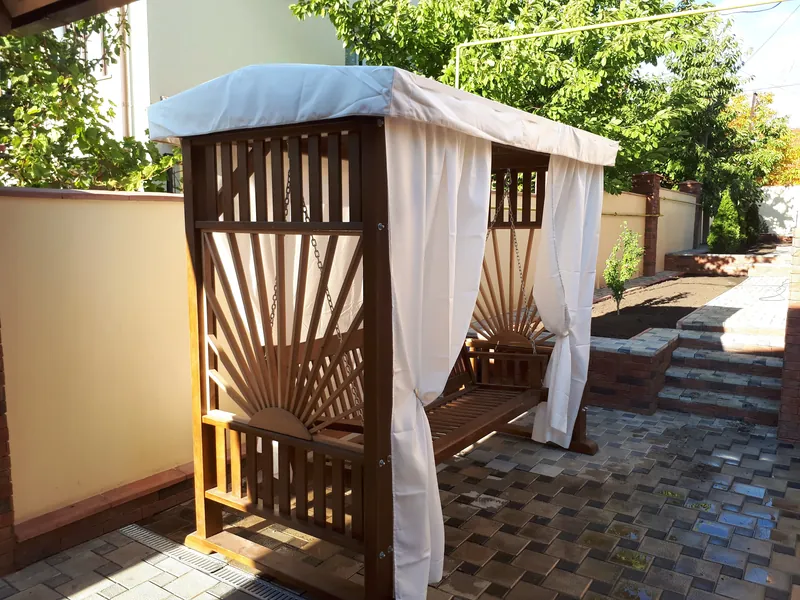 Leagăn din lemn cu panou lateral în evantai, copertină și perdele albe, în curte pavată — LEMNER.MD