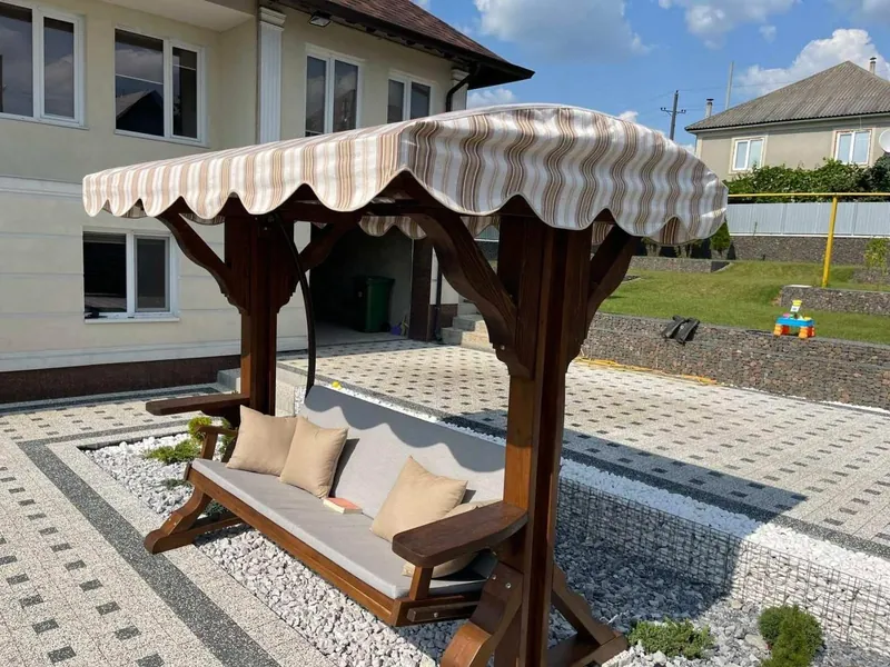 Leagăn din lemn tip foișor, cu copertină în dungi și perne crem, în curte amenajată cu pietriș — LEMNER.MD